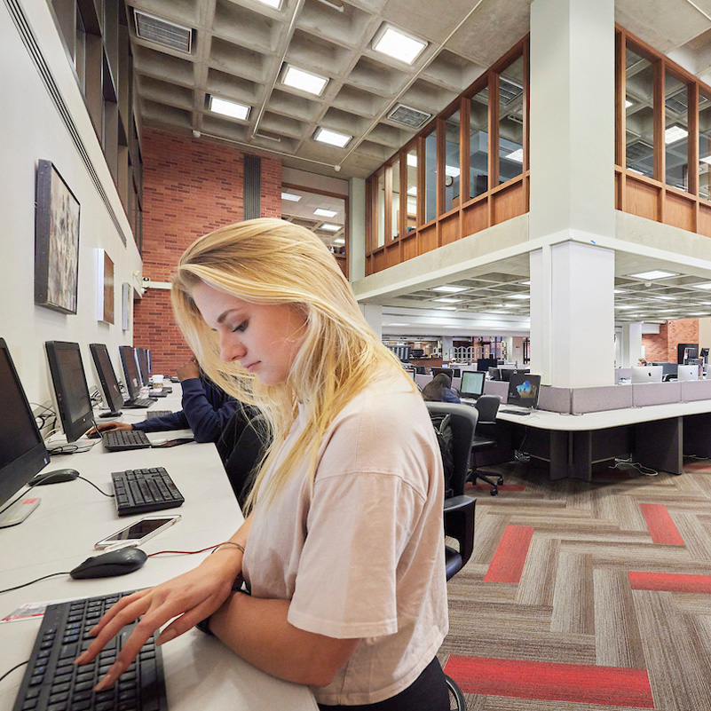 Young woman typing on a computer in a modern library, surrounded by sleek workstations and vibrant red and gray carpet.