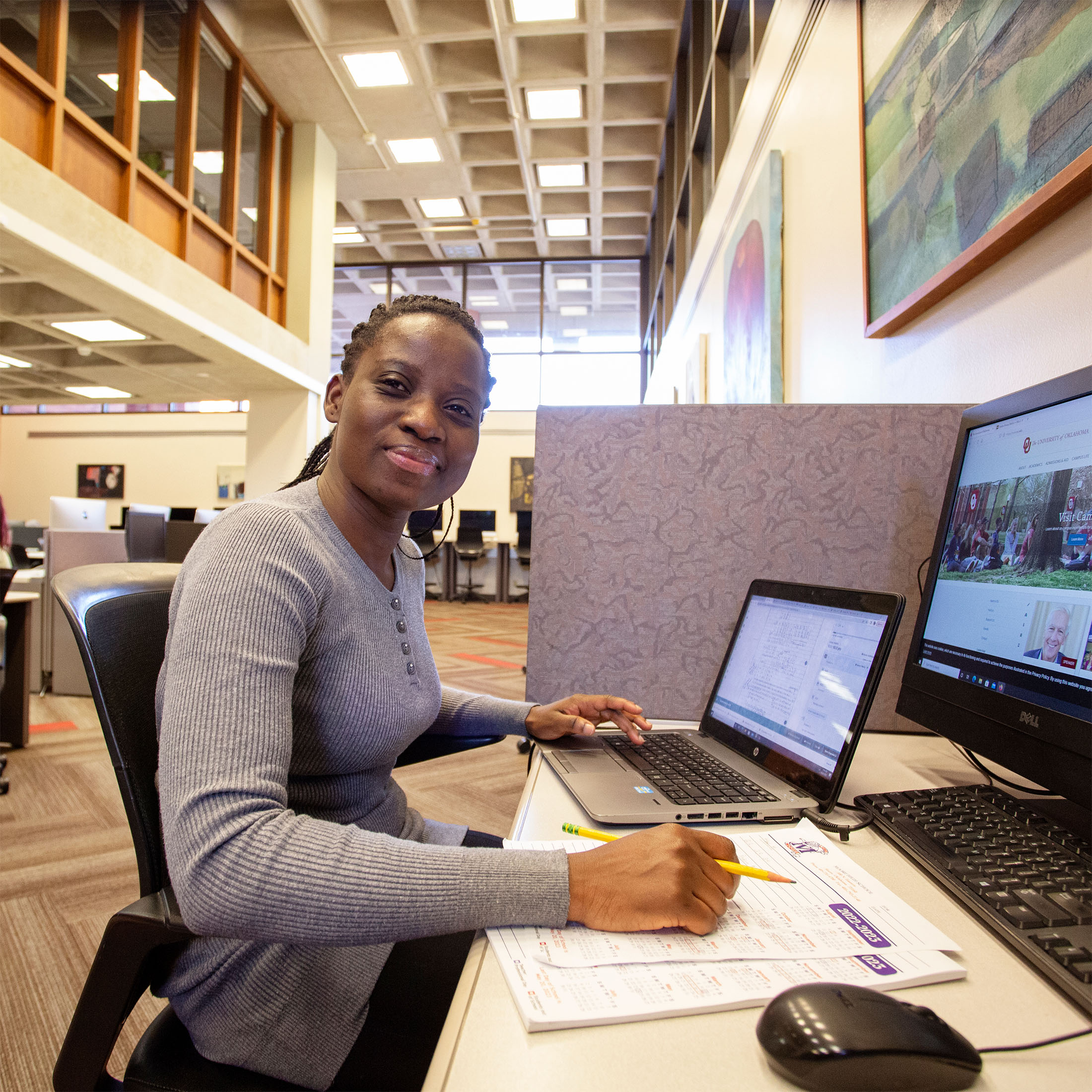 A woman sits at a desk in a library, smiling slightly at the camera while working on a laptop. Papers and a desktop computer are also on the desk. The environment is spacious and well-lit.