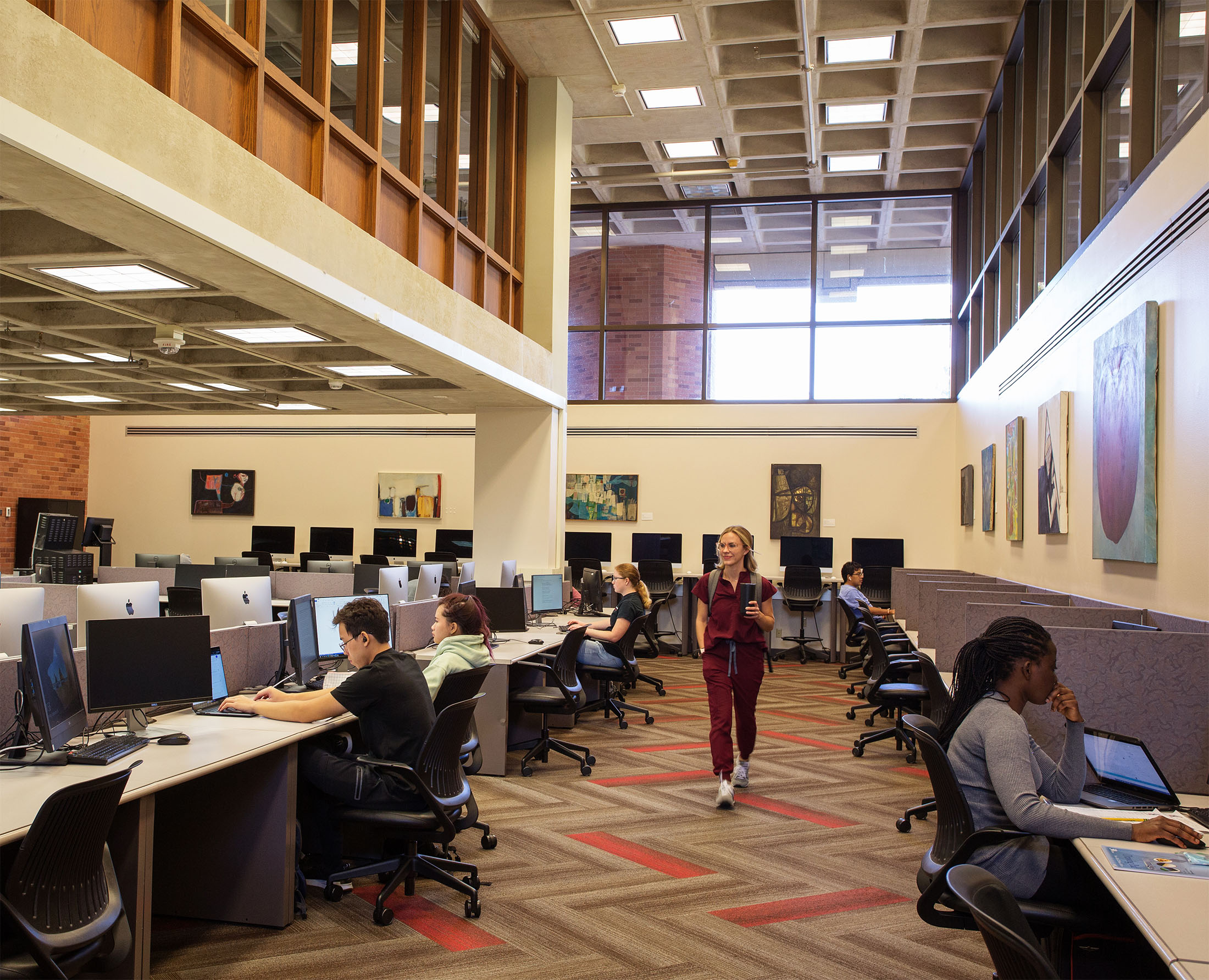 A modern library computer lab with people working at computer workstations on long tables. Art adorns the walls, and a person in red walks through, creating a studious atmosphere.