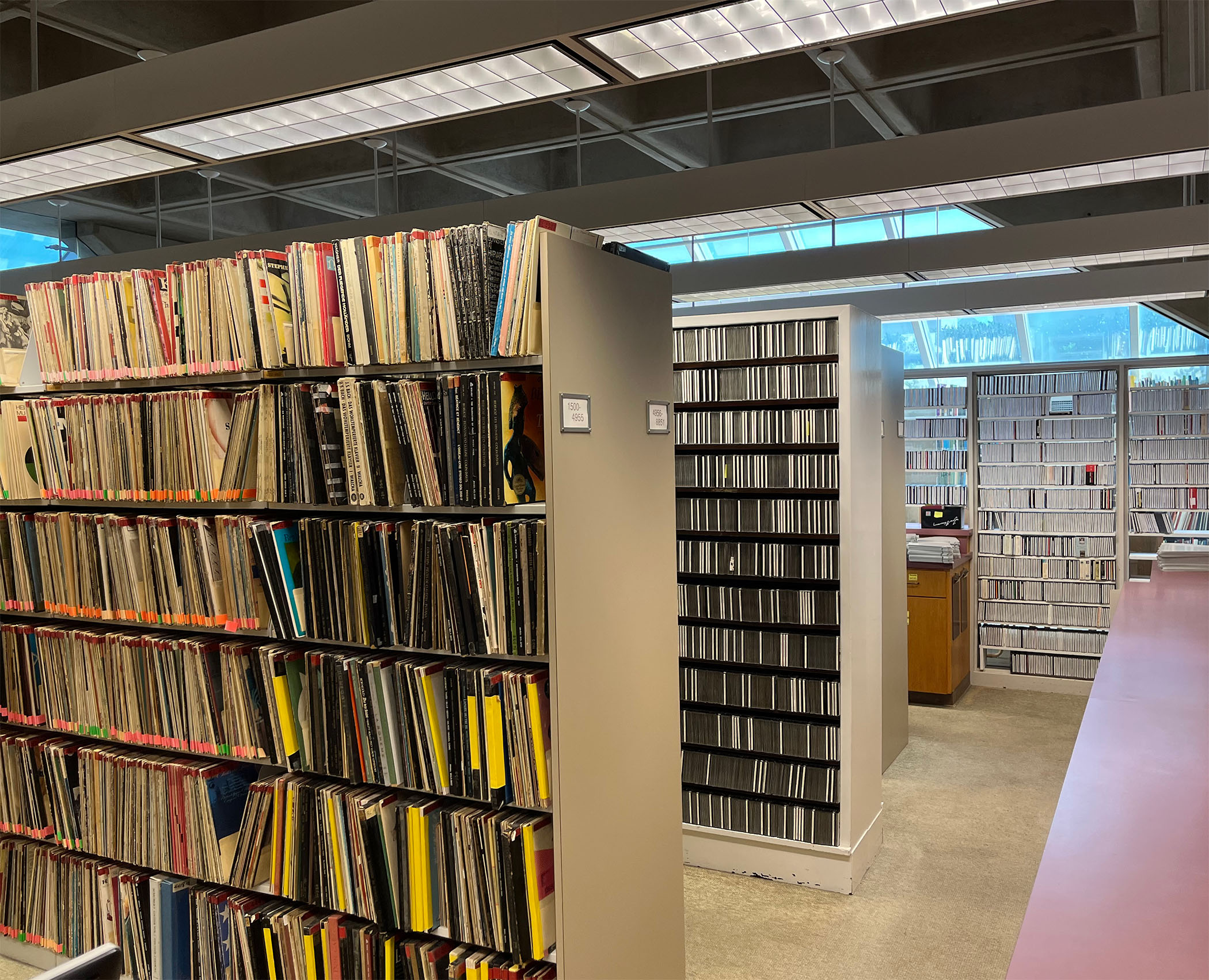 Library special collection archive with tall shelves filled with vinyl records and CDs, under bright fluorescent lighting. The space feels organized and quiet.