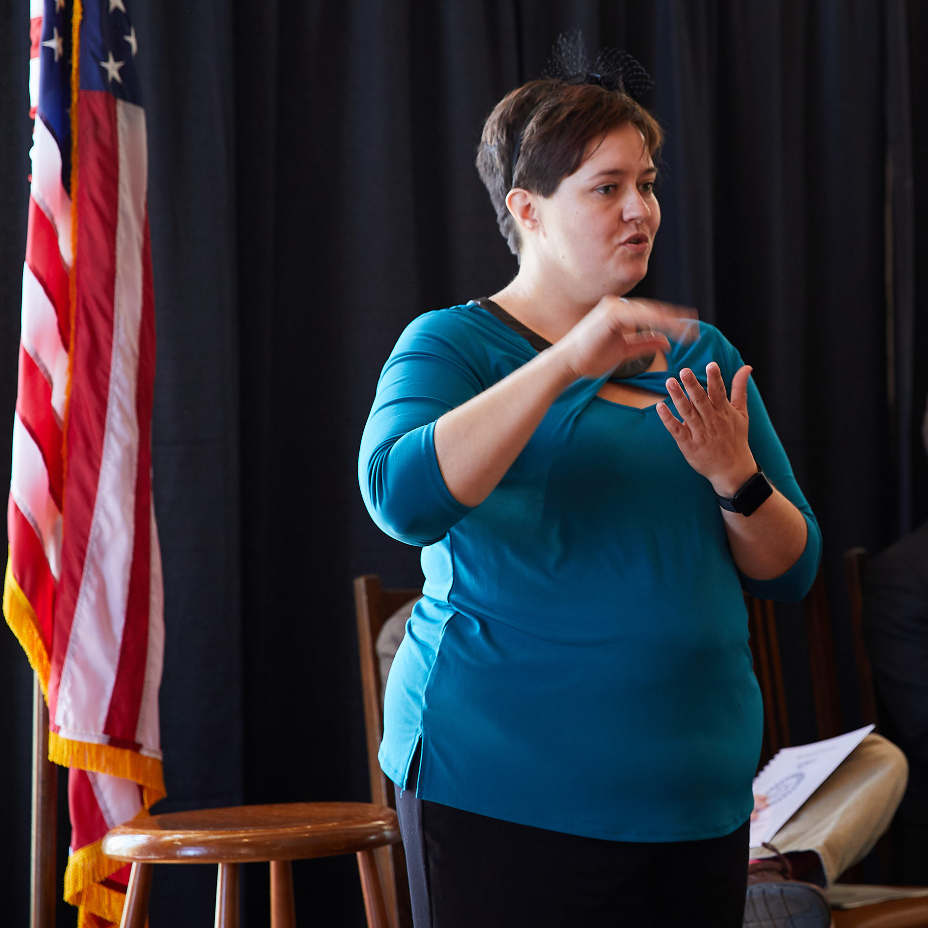 A woman in a blue top uses sign language to interpret a presentation being held in an auditorium. She is wearing a blue top and stands in front of a U.S. flag and black curtain.