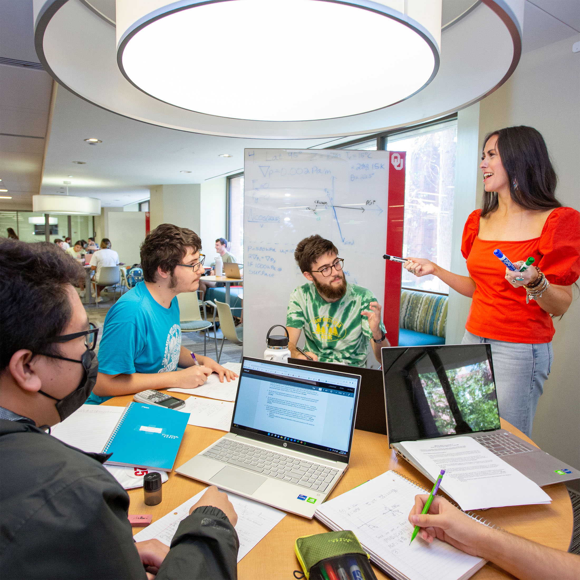 A woman stands by a whiteboard, smiling and gesturing with markers, as she gives a presentation to a group of four students. They sit and collaborate at a round table, with two laptops open, discussing concepts.