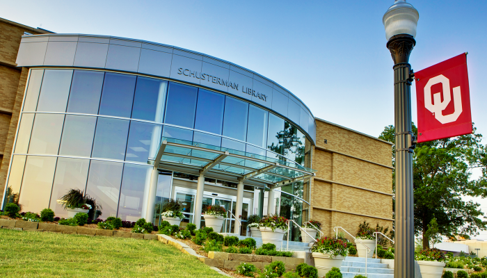 A modern glass-fronted library entrance with "Schusterman Library" sign on the top, front of the building.