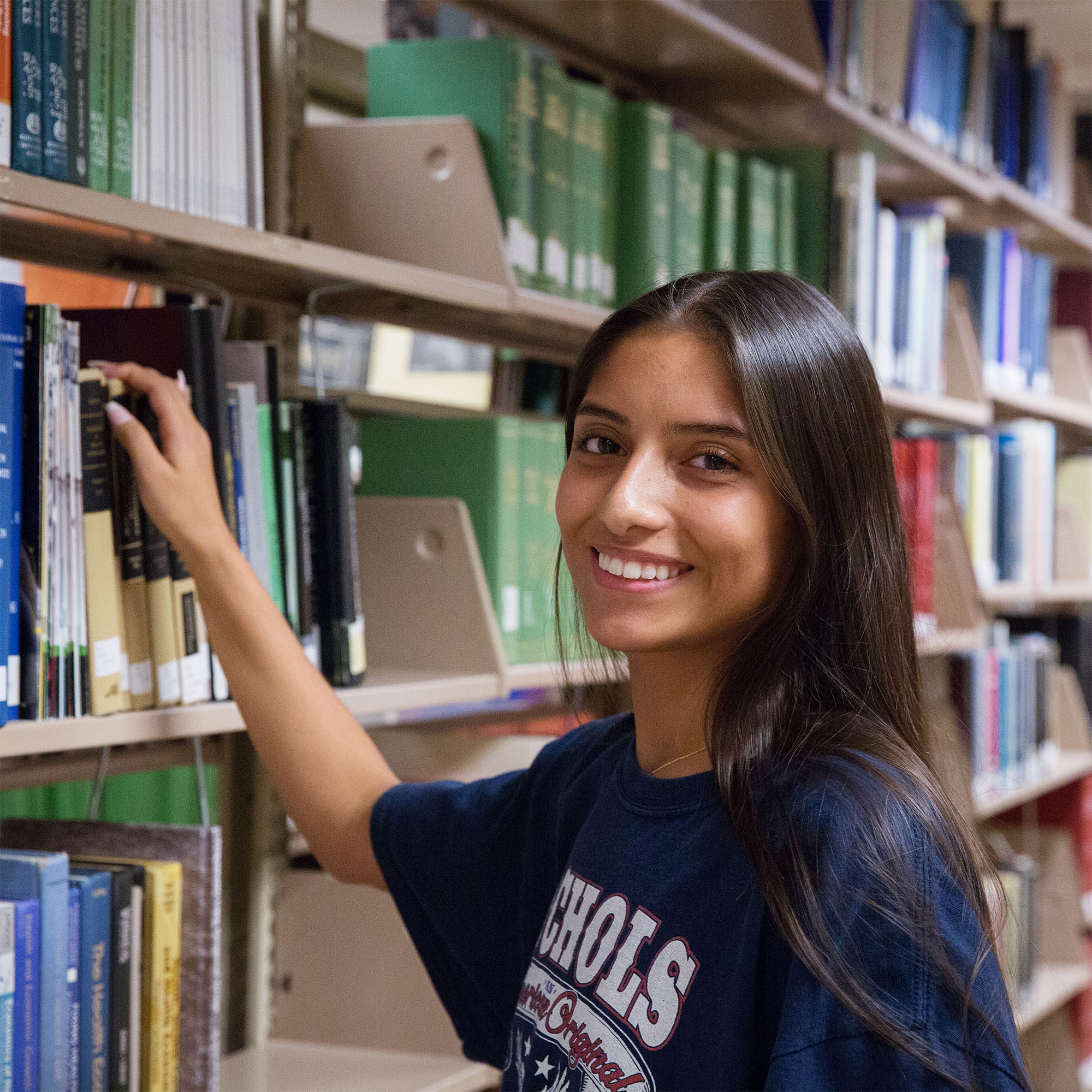 Smiling woman in a library, reaching for a book on a shelf. She's wearing a blue shirt. The atmosphere is calm and studious.
