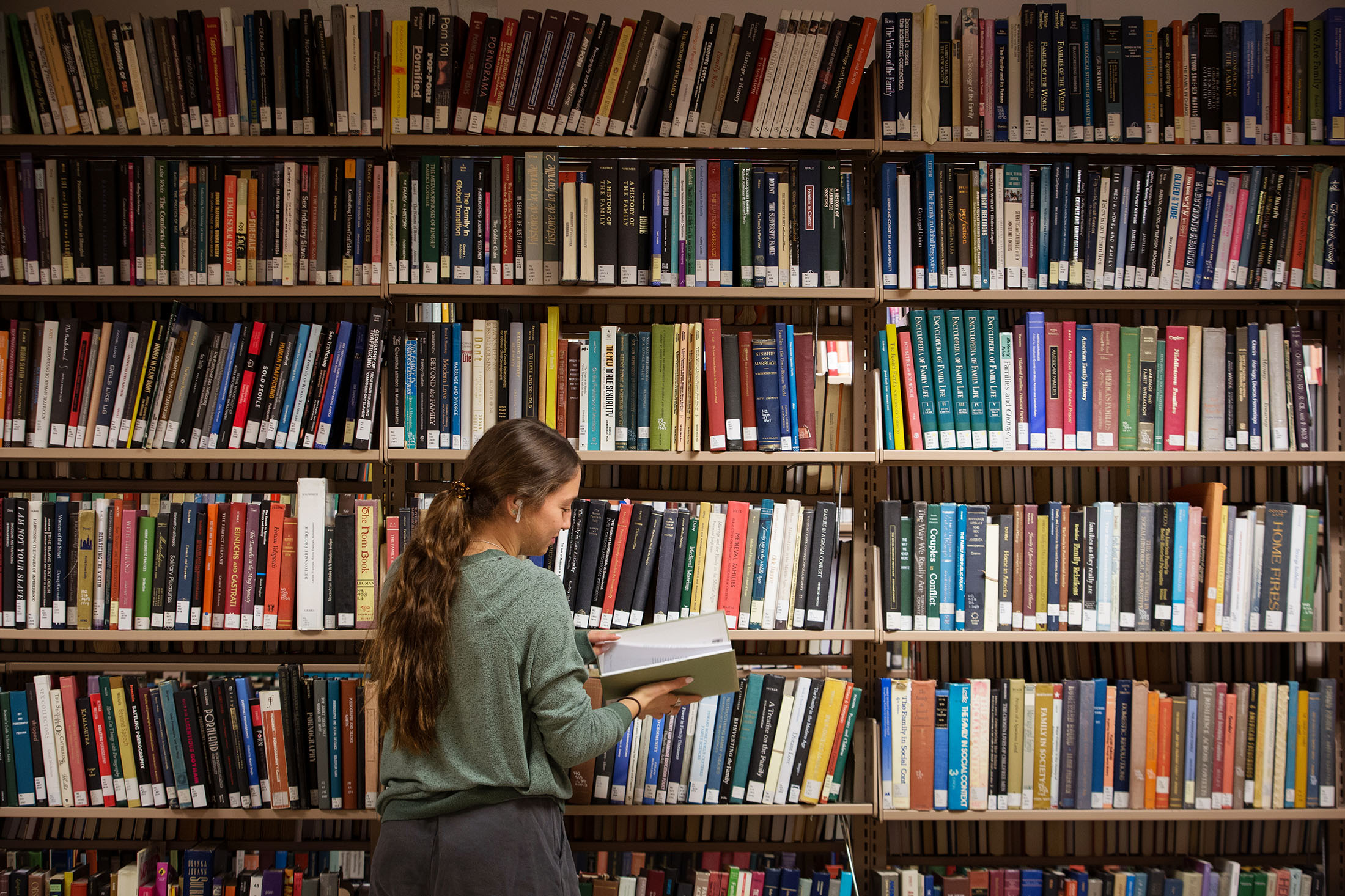 A woman with long hair, green sweatshirt and black pants stands reading a book in a library filled with colorful books on wooden shelves.
