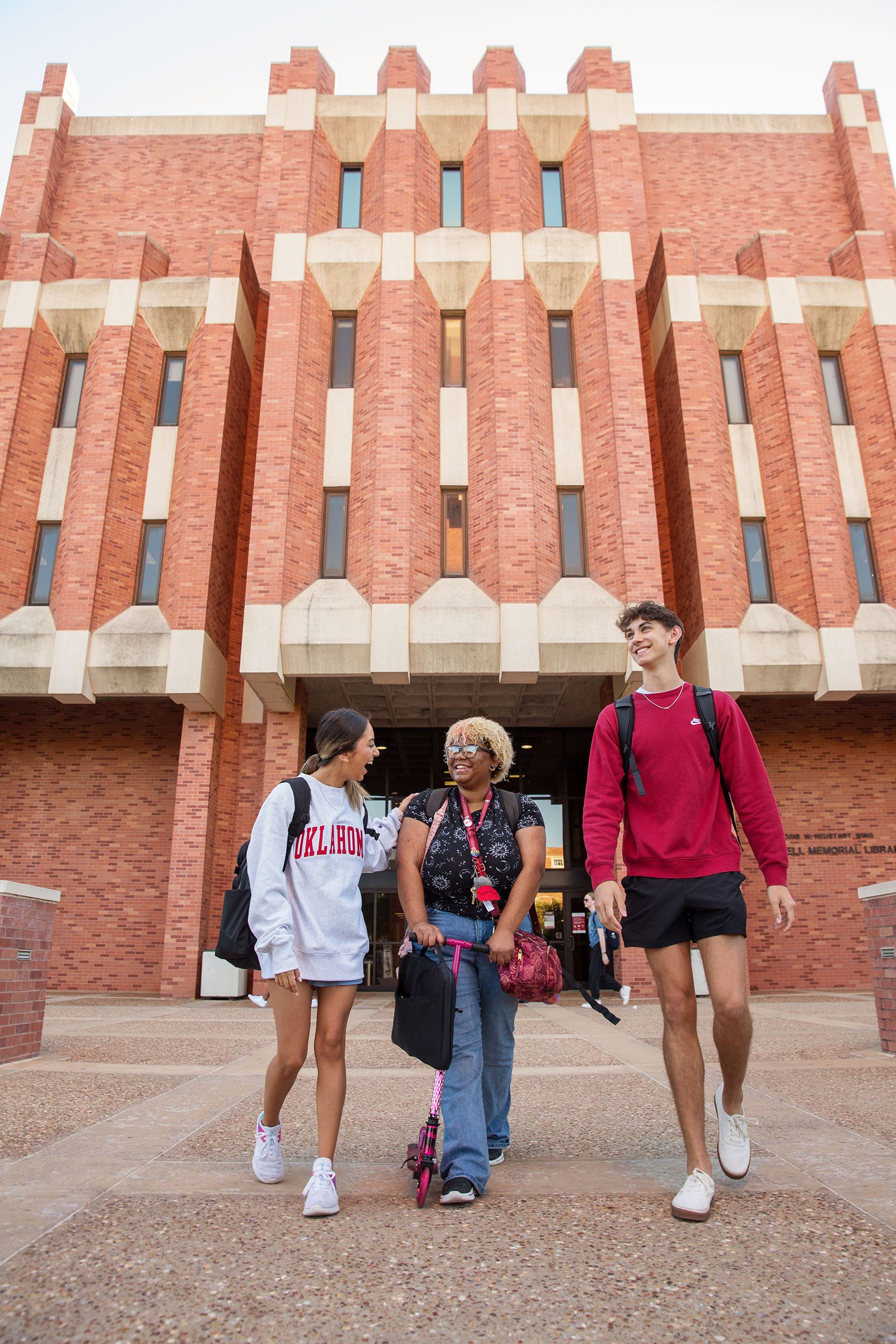Three students are walking joyfully in front of the west entrance to Bizzell Memorial Library, which features a red, brick facade and tall windows. The students are carrying backpacks and the central student pushes a pink scooter.