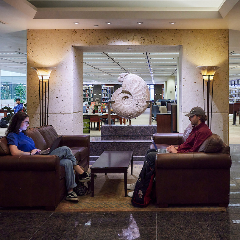 Two students sit on brown leather couches with laptops in the Youngblood Energy Library, framed by a large nautilus sculpture in the background. The ambiance is calm and studious.