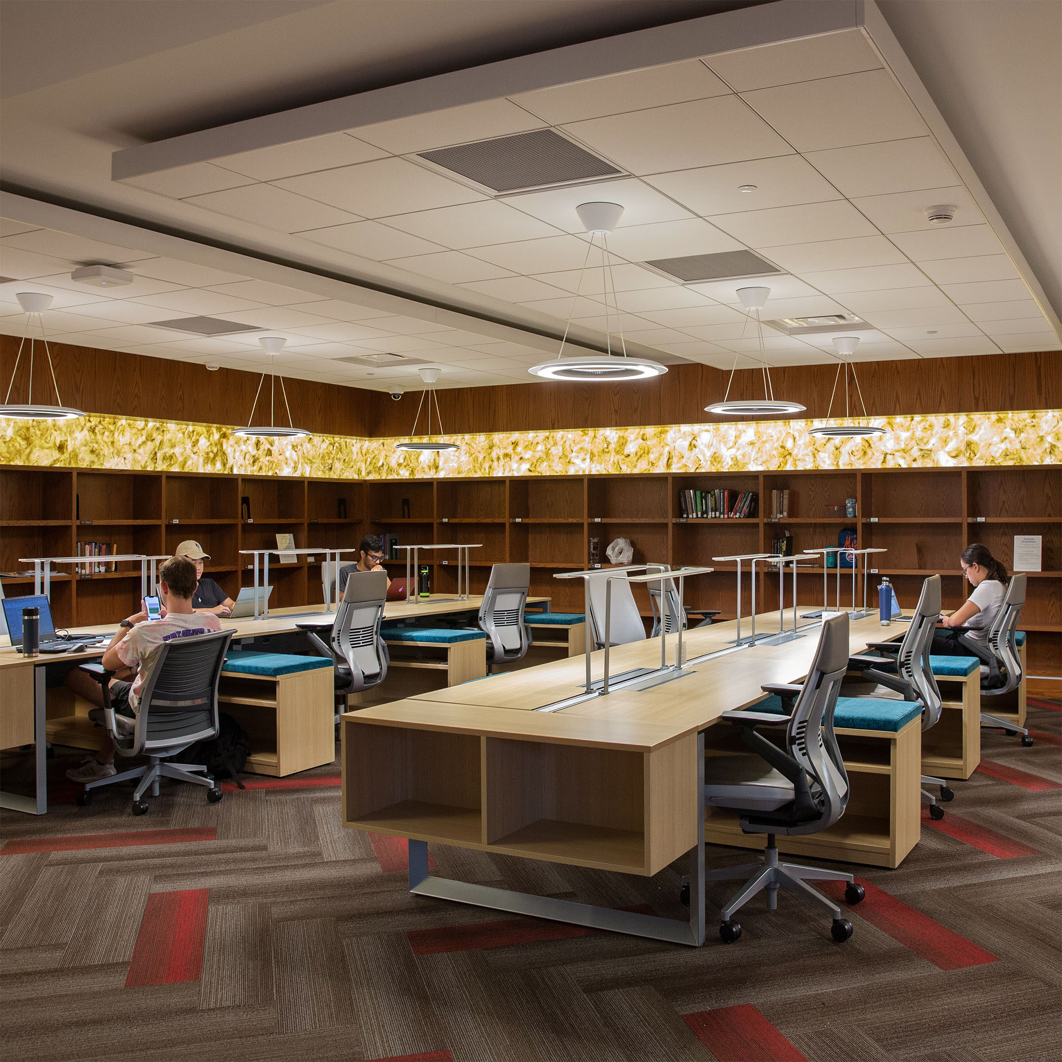 Modern library interior with long, wooden desks, open shelves, and ring-shaped pendant lights. Students seated, focused on laptops, creating a studious atmosphere.