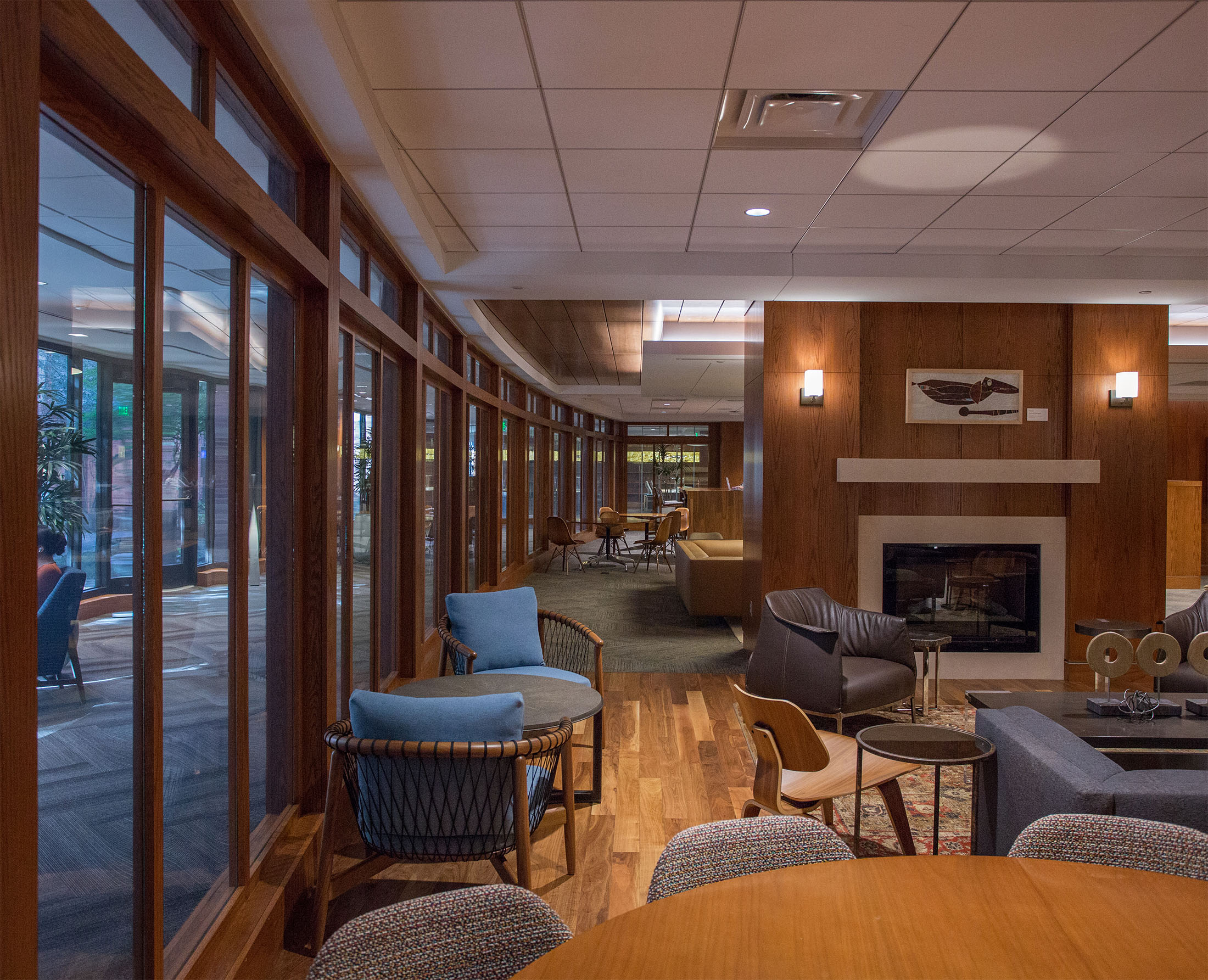 Cozy library lounge with wooden floors and walls, featuring a central fireplace, plush chairs, and large windows. Warm lighting creates an inviting atmosphere.