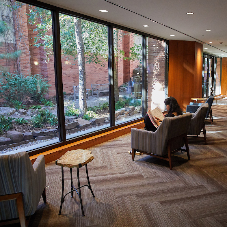 A woman reads in a cozy seating area with large windows showcasing a peaceful garden. The scene conveys tranquility and warmth with natural light.