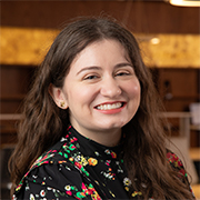 Morgan Briles smiles towards the camera. She has wavy brown hair and is wearing a floral blouse. She conveys a cheerful demeanor, set against a warm, blurred indoor background.
