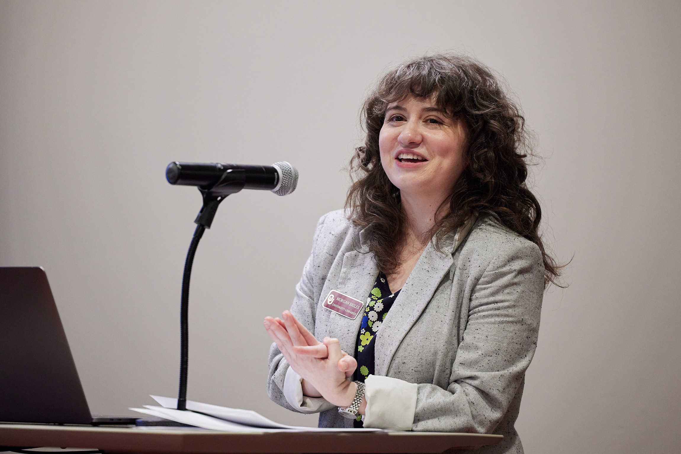 Morgan Briles speaks at a podium with a microphone. She has brown, curly hair and is wearing a gray blazer. She appears confident and engaging, conveying a positive tone.
