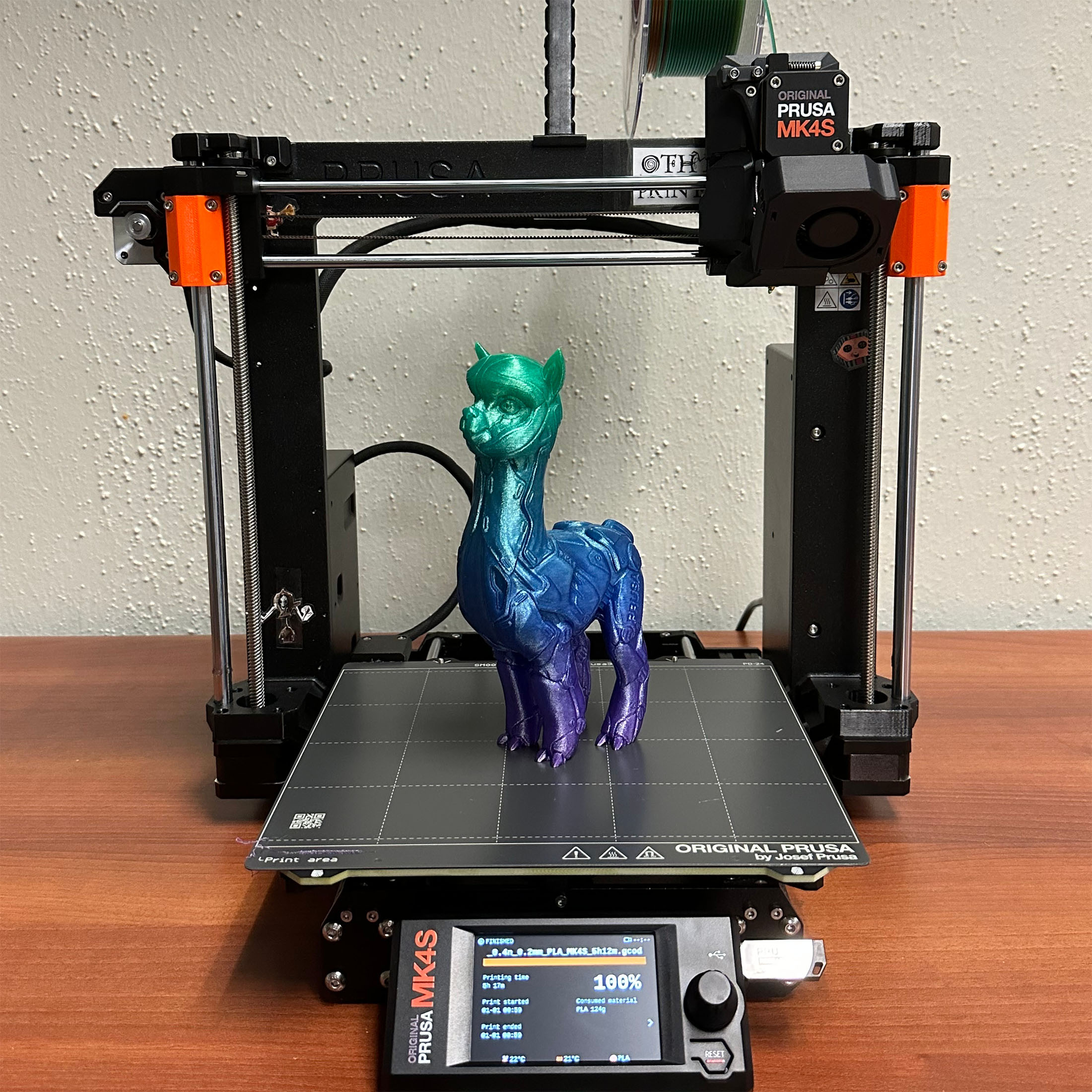 A colorful 3D-printed llama with a green head and blue-purple body on a Prusa MK4S 3D printer bed, set on a wooden table.