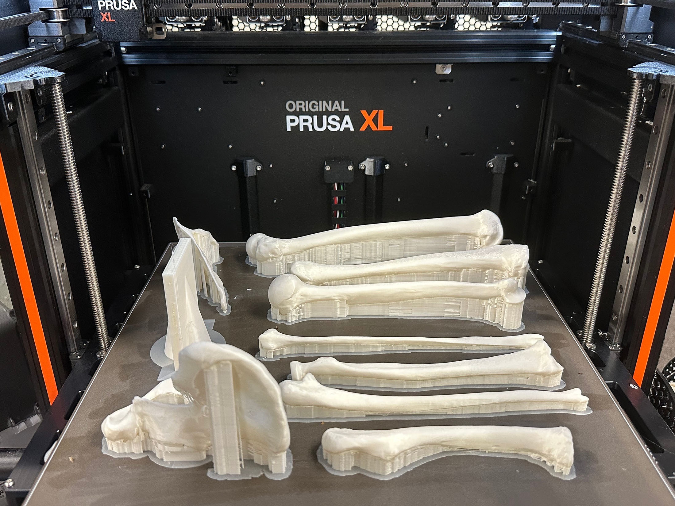 A Prusa XL 3D printer displays several white, 3D-printed human bones on its platform.