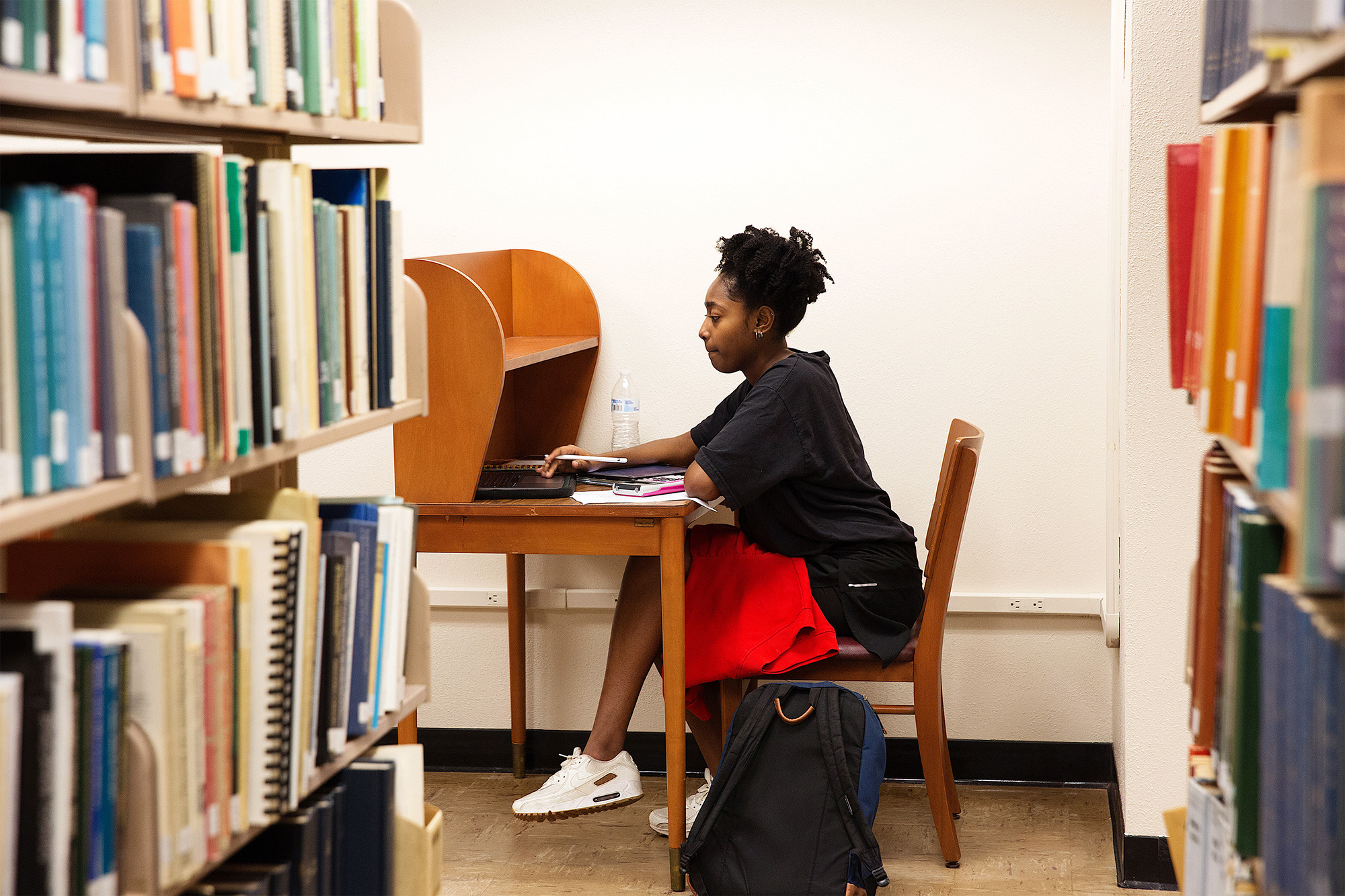 A person is sitting at a wooden desk in a library, focused on a laptop. Bookshelves surround them. They appear engaged, creating a studious atmosphere.