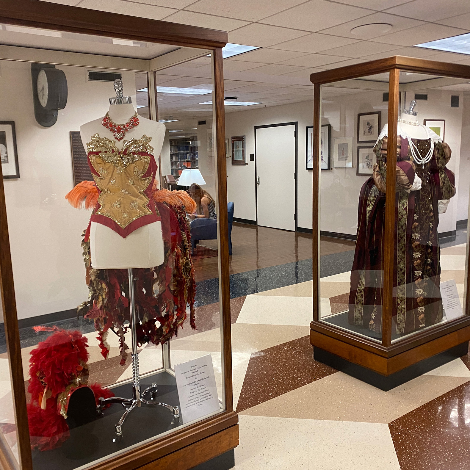 In the hallway of Bizzell Memorial Library, two glass cases with mannequins wearing costumes are on display. One costume has a gold and red bodice with feathers; the other, a vintage gown with drapes, pearls.