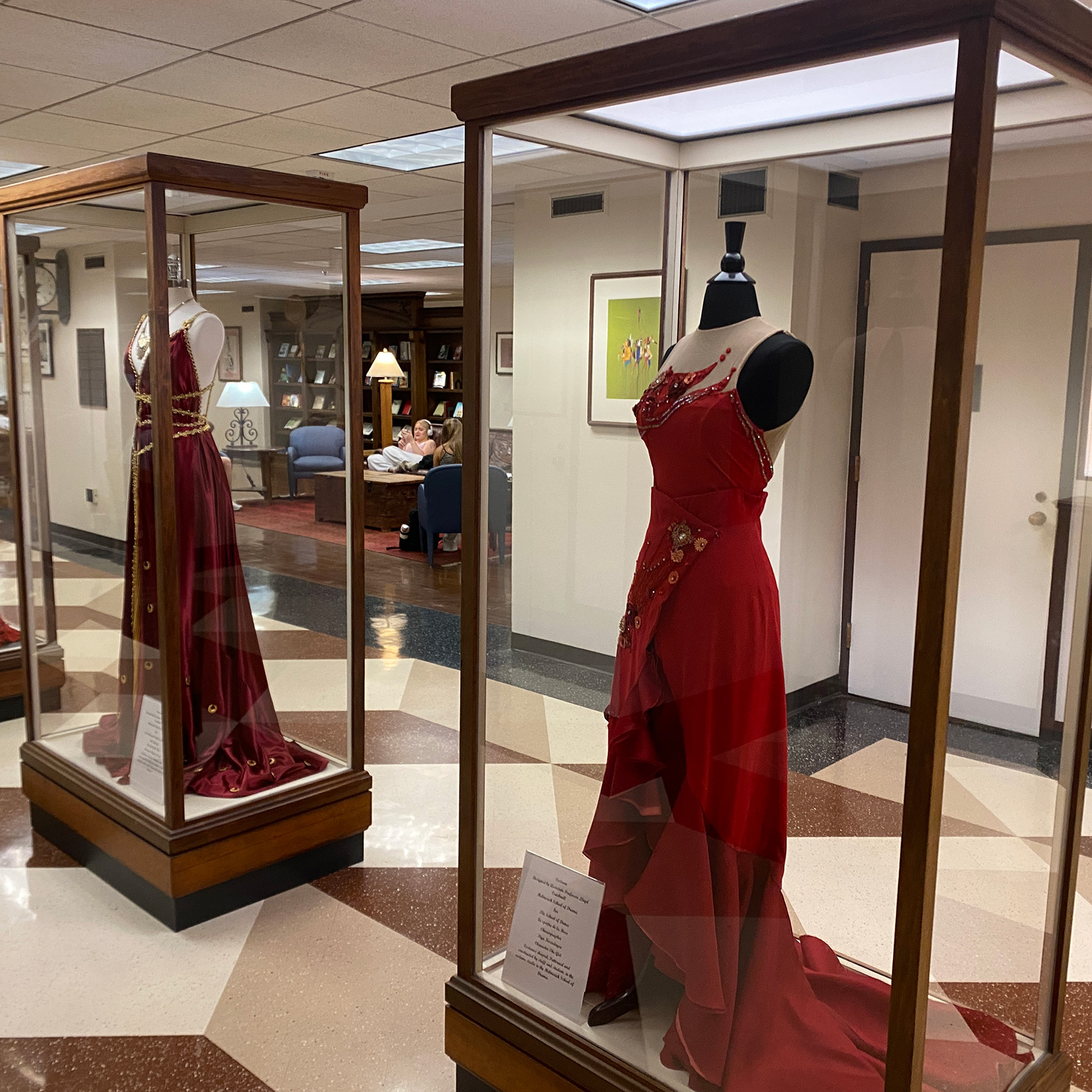 In the hallway of Bizzell Memorial Library, two glass cases with mannequins dressed in costumes are on display. One costume is a red evening gown adorned with decorative elements is displayed on a mannequin inside a glass case.
