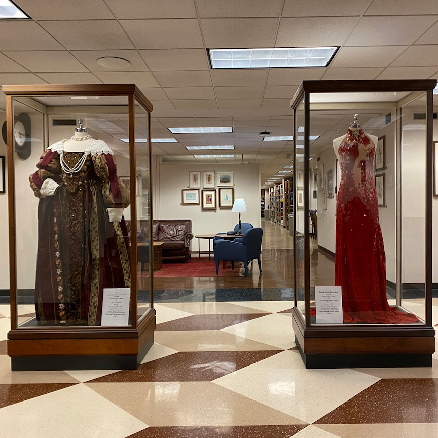 In the hallway of Bizzell Memorial Library, two glass cases with mannequins dressed in costumes are on display. The left case holds a Renaissance-style dress, while the right features a glittering red gown.