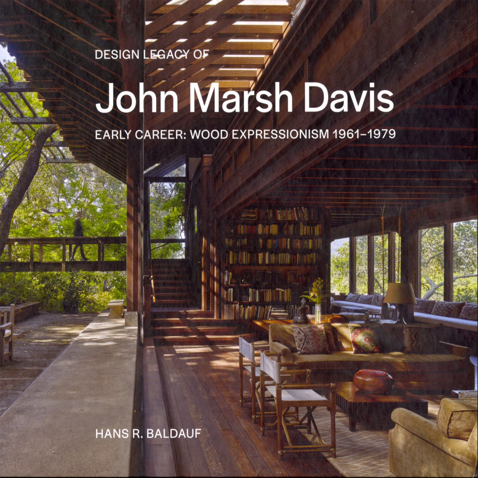 Book cover title "Design Legacy of John Marsh Davis, Early Career: Wood Expressionism 1961–1979" by Hans R. Baldauf. The white text overlays a background photo of a Luxurious wooden interior with large windows, sunlight filtering through a trellis. Cozy seating with cushions, bookshelves, and forest views.
