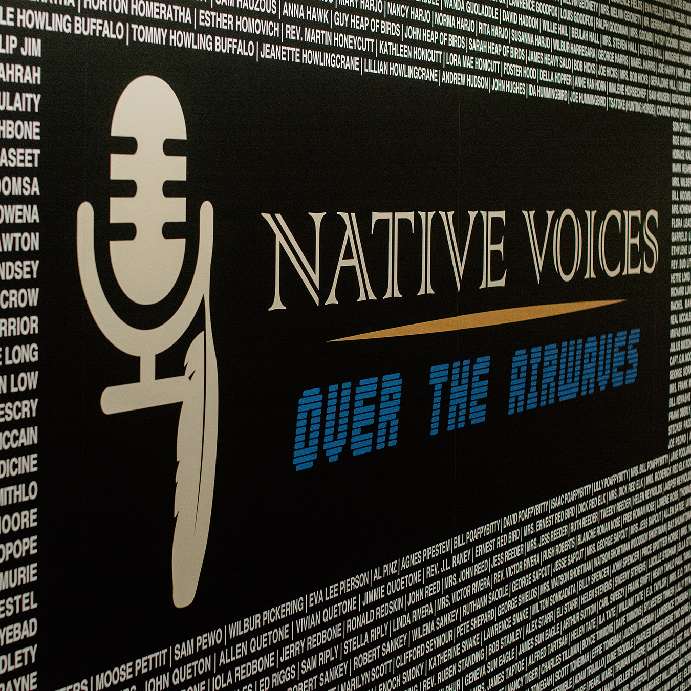 Image featuring a large microphone graphic with a feather, alongside text "Native Voices Over the Airwaves" in bold white and blue letters on a black rectangular shape. Surrounding background text lists numerous names in white on a black background.