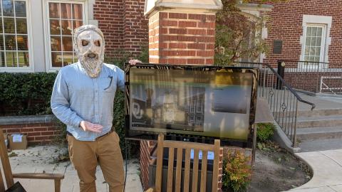 Outside a red, brick building, a person wearing a creepy mask stands beside a digital screen on a stand. The screen is displaying OU architectural images.
