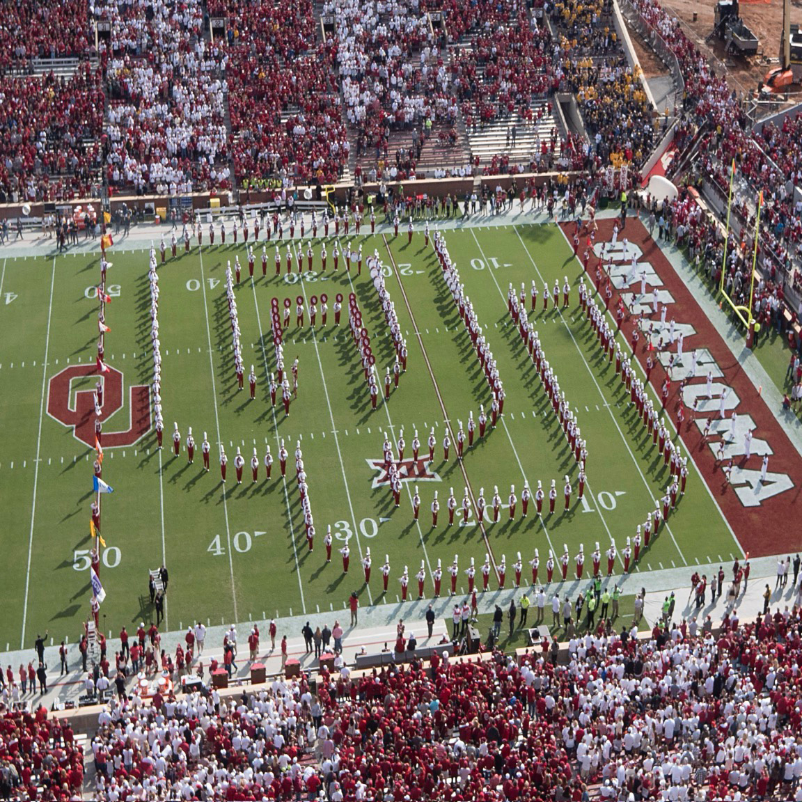 The Pride of Oklahoma marching band forms a large "OU" shape on the OU football field, filled with cheering crowds. The field is bordered by "OKLAHOMA" in bold, white letters on a crimson background.