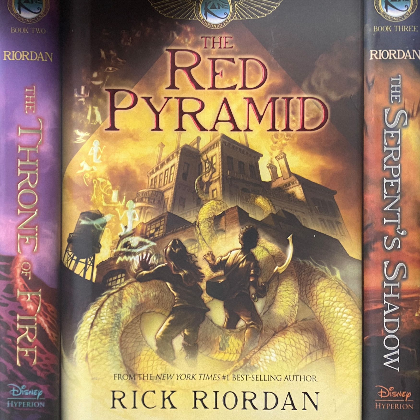 The image displays three books by Rick Riordan. In the center, a book cover of "The Red Pyramid" features two figures facing a large snake with an ancient building in flames behind them, evoking adventure and mystery. To the left is the spine of "The Throne of Fire" and to the right is the spine of "The Serpent's Shadow."