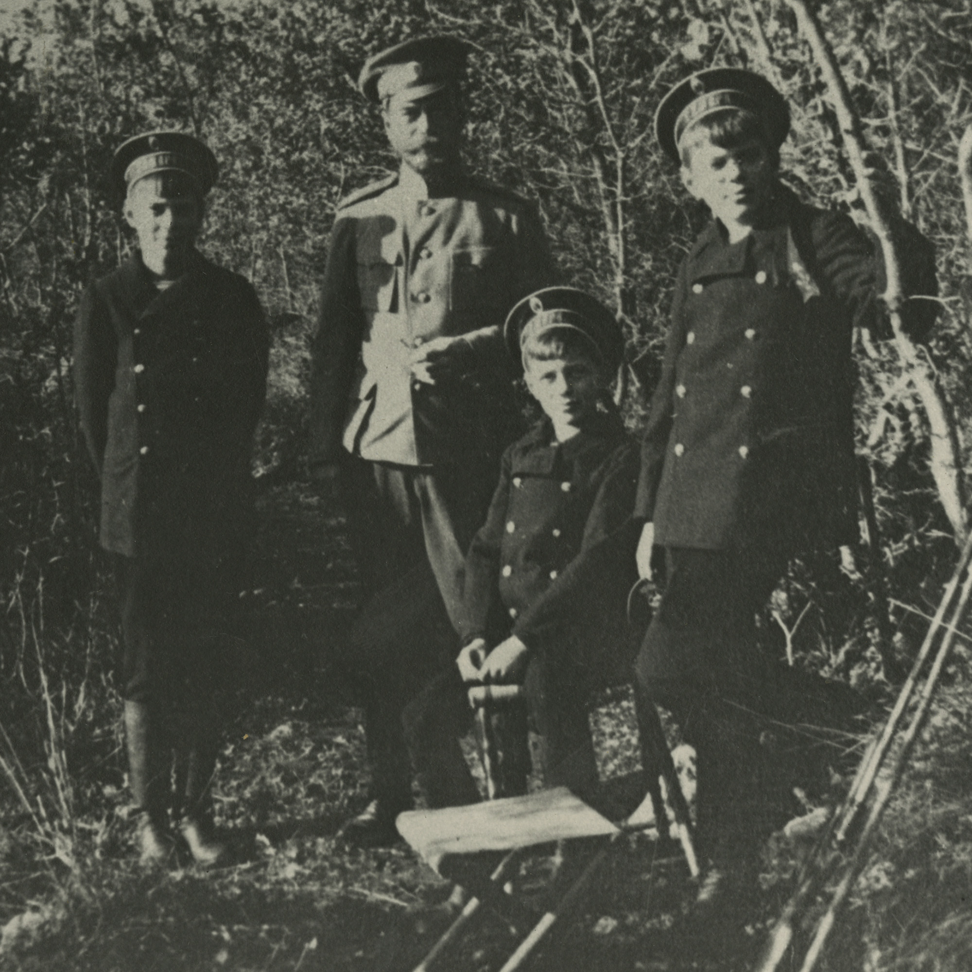 Black and white photo of four people outdoors, three young boys in uniform caps and coats, and one older in military attire, with a small, foldable stool in front.