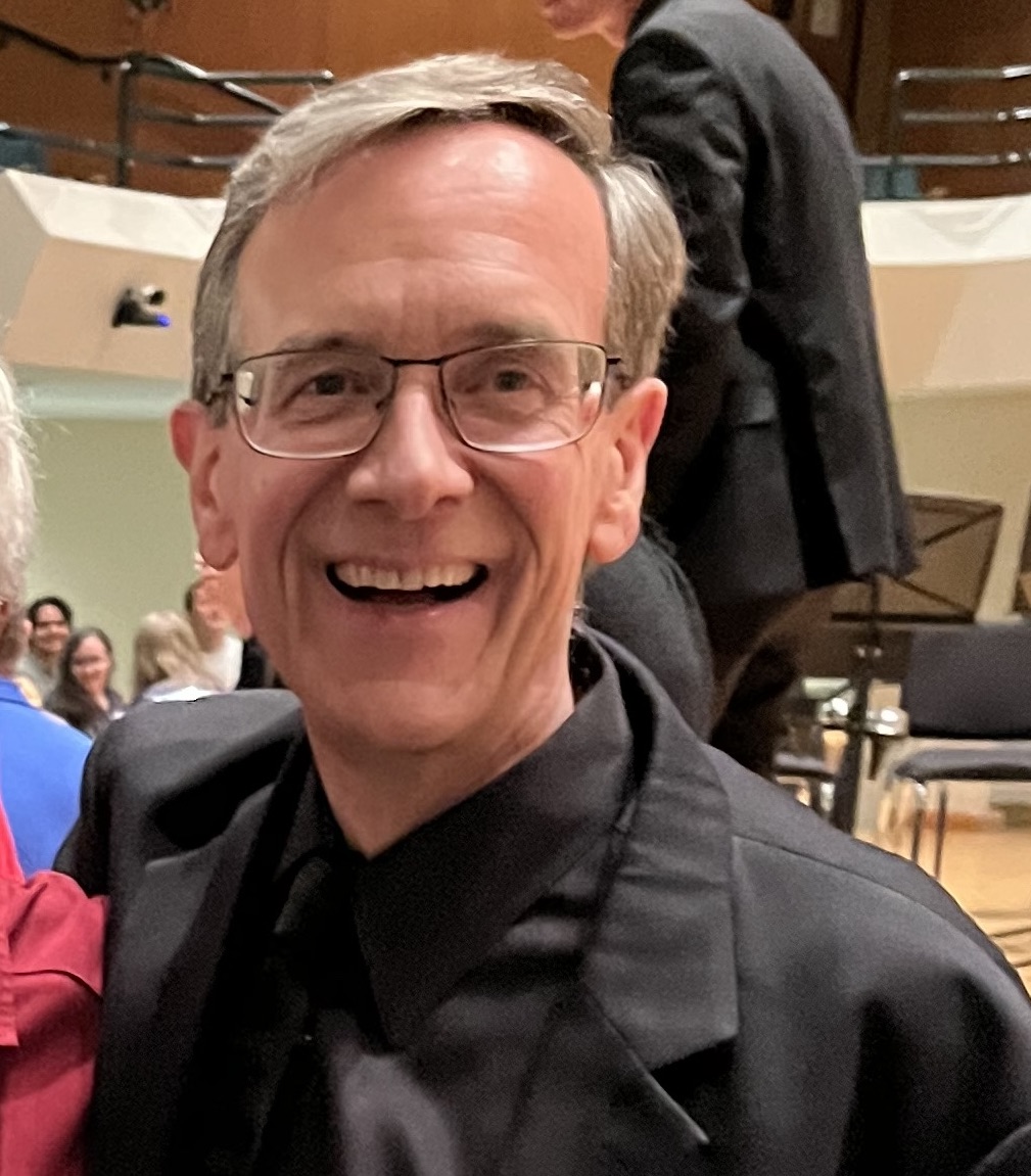 Matt Stock wears glasses, a black collared shirt, and a black blazer, smiling at the camera. He is attending an indoor musical performance in a multi-level auditorium. In the background, blurred people are seated facing a stage with music stands and chairs.