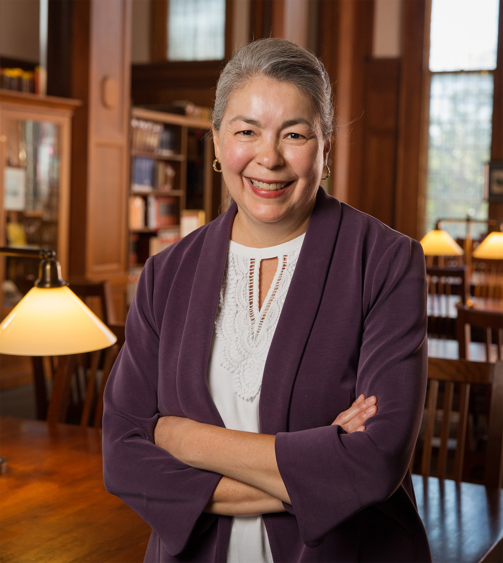 Lina Ortega has gray hair and is smiling towards the camera. She stands confidently in a library, her arms crossed, wearing a purple jacket and white blouse, with warmly lit lamps and bookshelves behind her.
